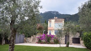 Une maison moderne à deux étages, dotée d’un escalier en pierre menant à l’entrée, entourée d’une végétation luxuriante et avec une chaîne de montagnes en toile de fond.