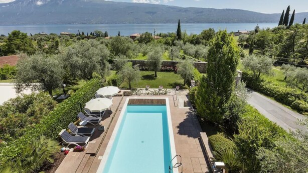 Une piscine rectangulaire bordée de chaises longues et d’ombrelles, entourée d’une végétation luxuriante et offrant une vue sur un lac et des montagnes.
