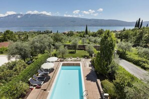 A rectangular swimming pool with lounge chairs and umbrellas, surrounded by lush greenery and a view of a lake and mountains.