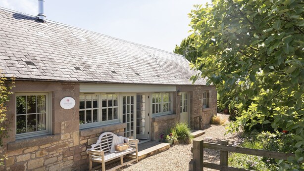 A stone cottage with a sloped roof, a bench outside, and a garden with a wooden fence. - HERITAGE COTTAGE set in peaceful rural surroundings, Scottish Borders (Duns)
