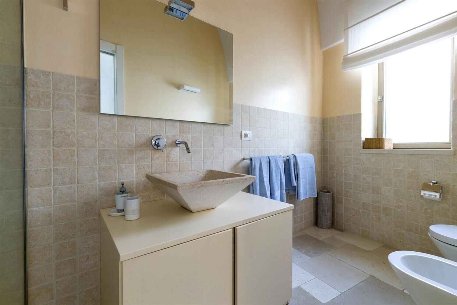 Un baño con pared de baldosas color crema, un lavamanos blanco con lavabo de piedra, un espejo sobre el lavamanos, una ventana con vistas y una bañera blanca.