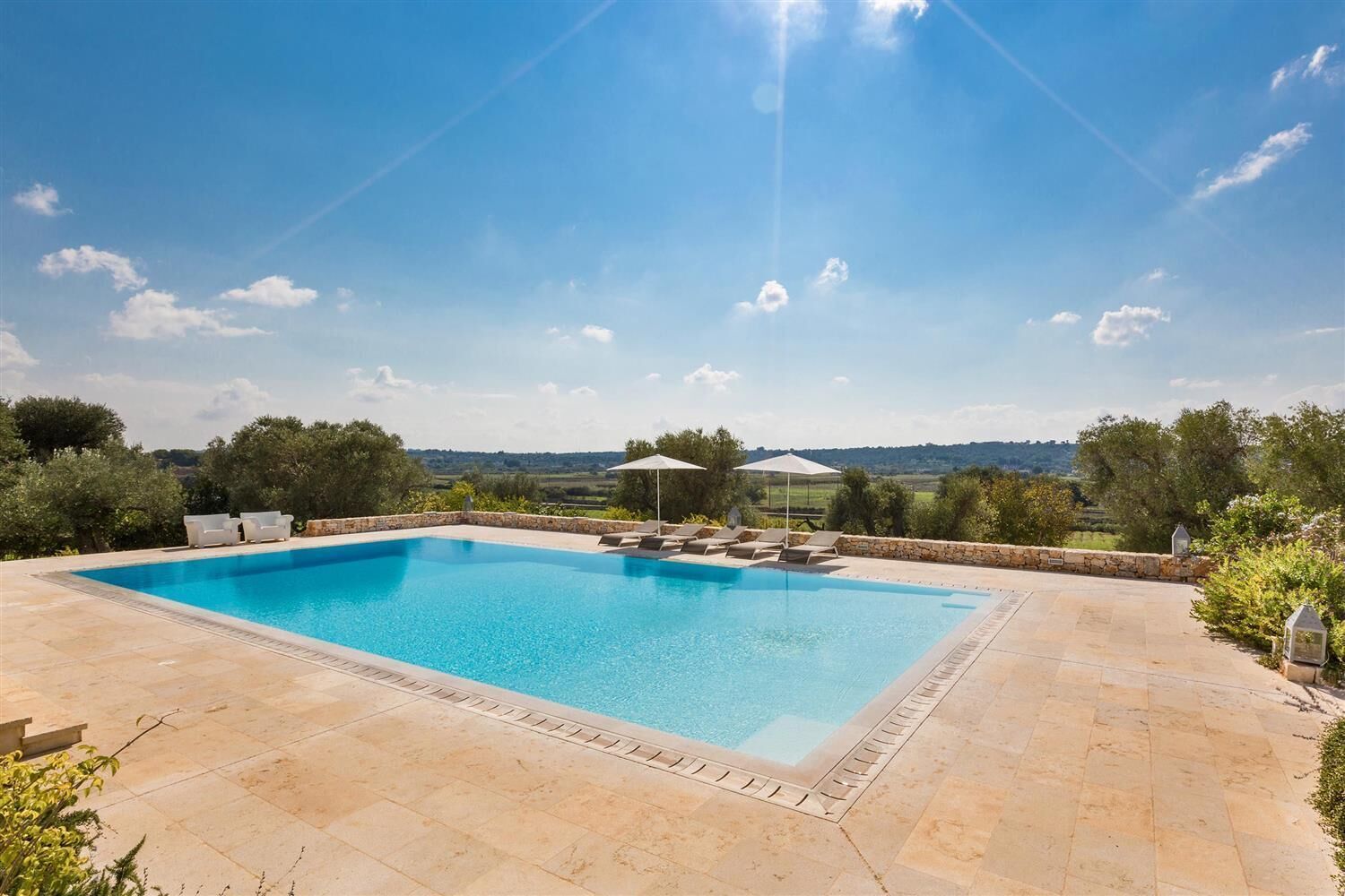 A rectangular swimming pool with a stone border, surrounded by a patio area with lounge chairs and umbrellas.