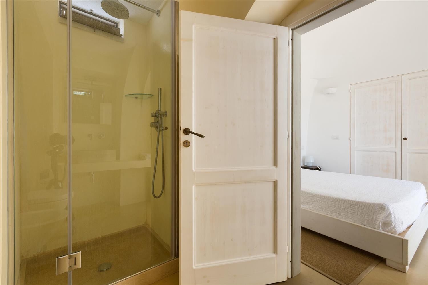 A room with a glass shower enclosure, a white door, and a bed with a white bedspread.