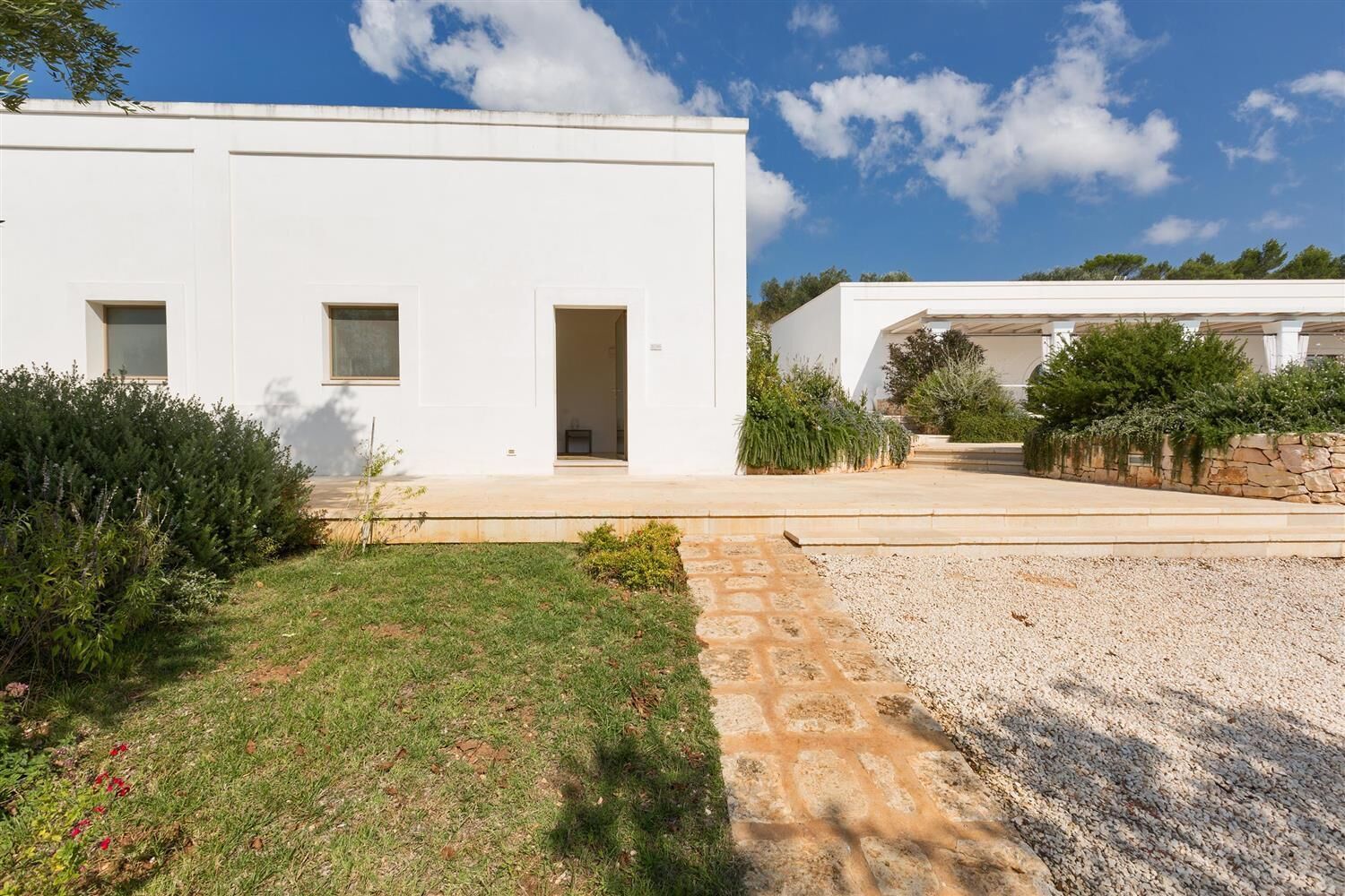 A modern white building with a paved driveway, a small garden area, and a gravel path.