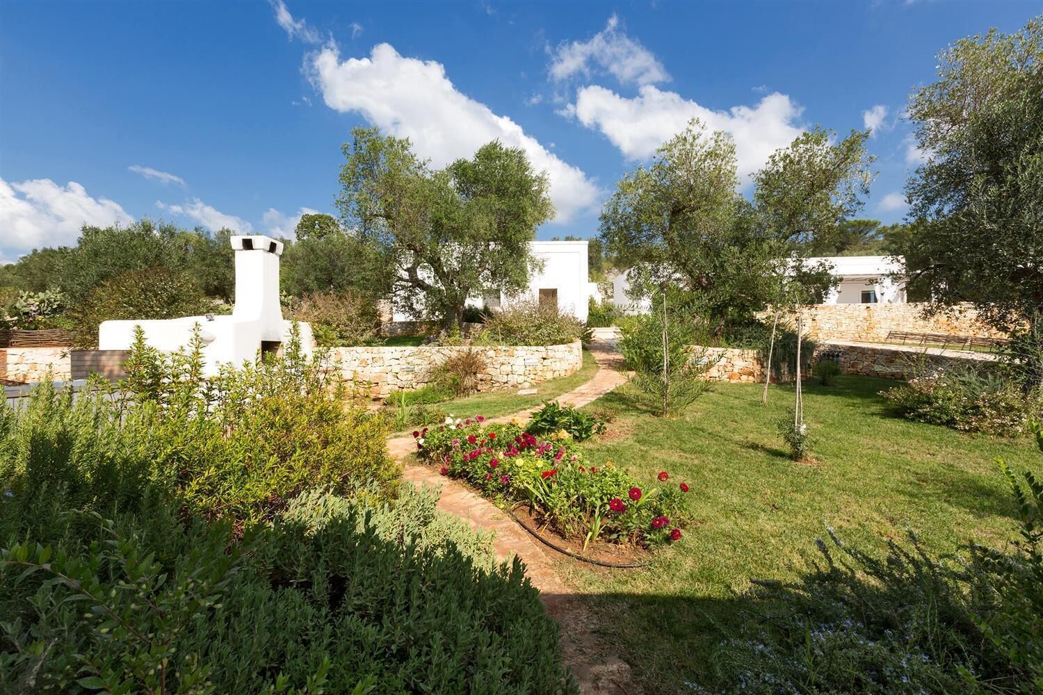 Un jardin avec un sentier menant à des bâtiments blancs, entouré de verdure et d’arbres.