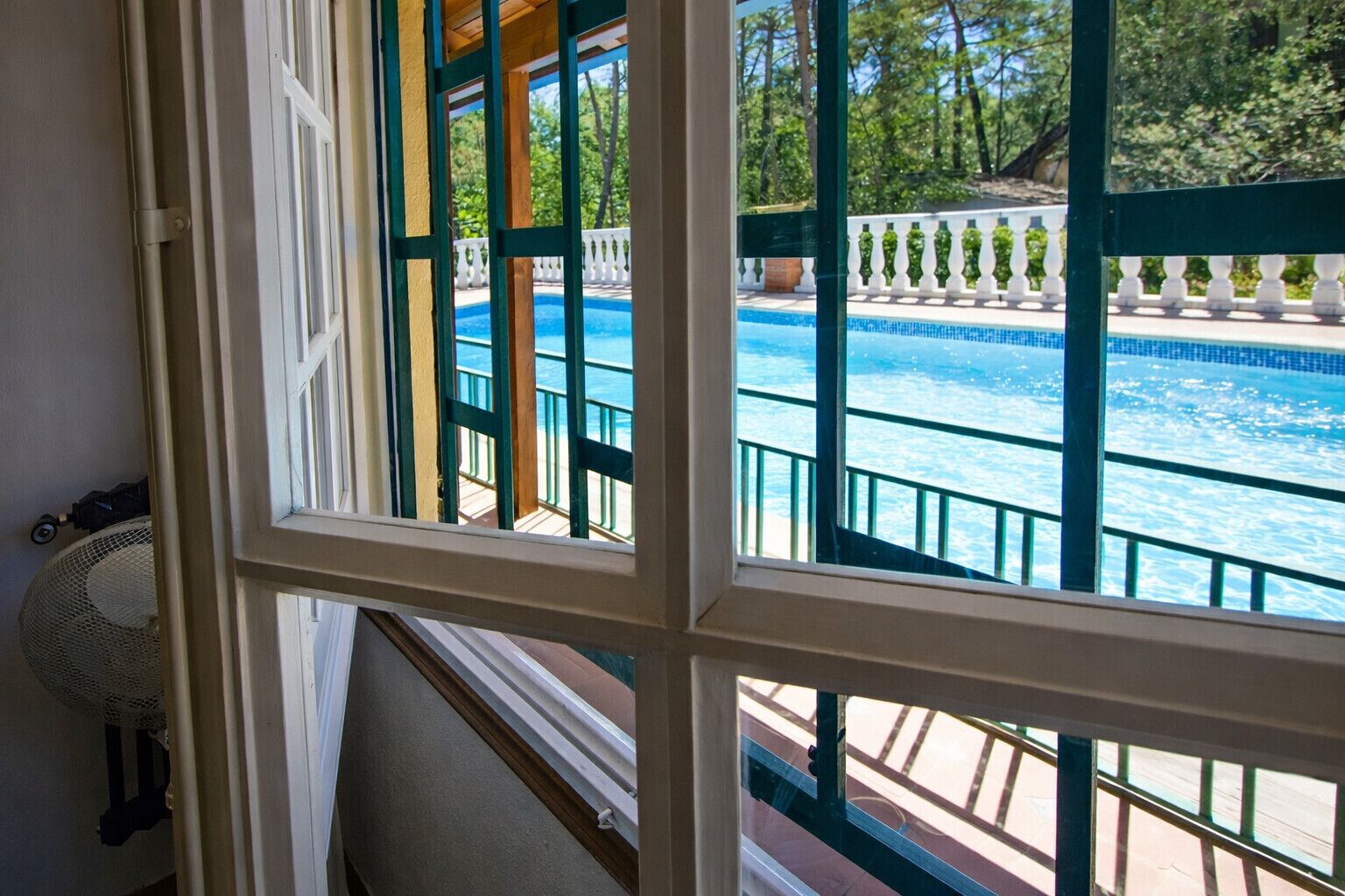 Vista de una piscina con cubierta de madera y barandilla blanca, visible a través de una ventana de marcos blancos y detalles verdes.