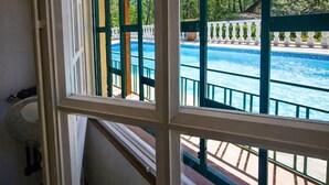 A view of a swimming pool with a wooden deck and white balustrade, seen through a window with white frames and green accents.