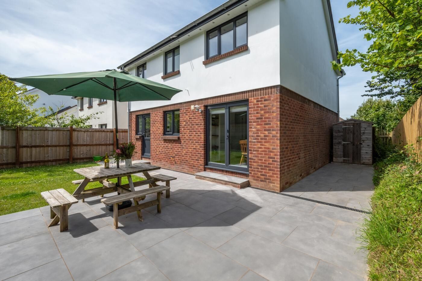 A modern two-story house with a patio area featuring a wooden table and benches, a green umbrella, and a brick exterior.