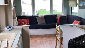 Un espacio habitable compacto con kitchenette, un sofá con cojines estampados, una mesa de comedor y vistas al exterior a través de grandes ventanales.