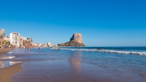 Une plage avec une formation rocheuse imposante qui se détache à l’horizon, un ciel dégagé et un paysage urbain avec des bâtiments qui bordent la côte.