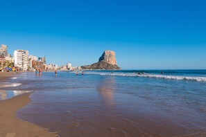 A beach with a prominent rocky formation in the distance, a clear sky, and a cityscape with buildings along the shore. - Apartment Brisa - PlusHolidays (Calpe)