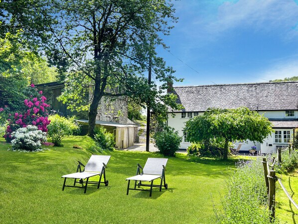 A garden with two lawn chairs, a stone building, and a white house with a sloped roof.