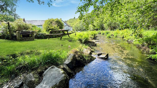 En bänk av sten, ett picknickbord, en klar bäck och ett hus i bakgrunden.