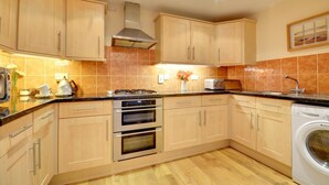A modern kitchen with wooden cabinets, a gas stove, an oven, a toaster, and a washing machine. - 2 Bed in Woolacombe (oc-narr9) (Woolacombe)