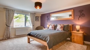 A bedroom with a bed, a bedside table with a lamp, a window with curtains, a radiator, and a mirror on the wall. - 2 Bed in Woolacombe (oc-narr9) (Woolacombe)