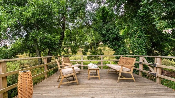 A wooden deck with two wicker chairs and a small table, surrounded by lush greenery.
