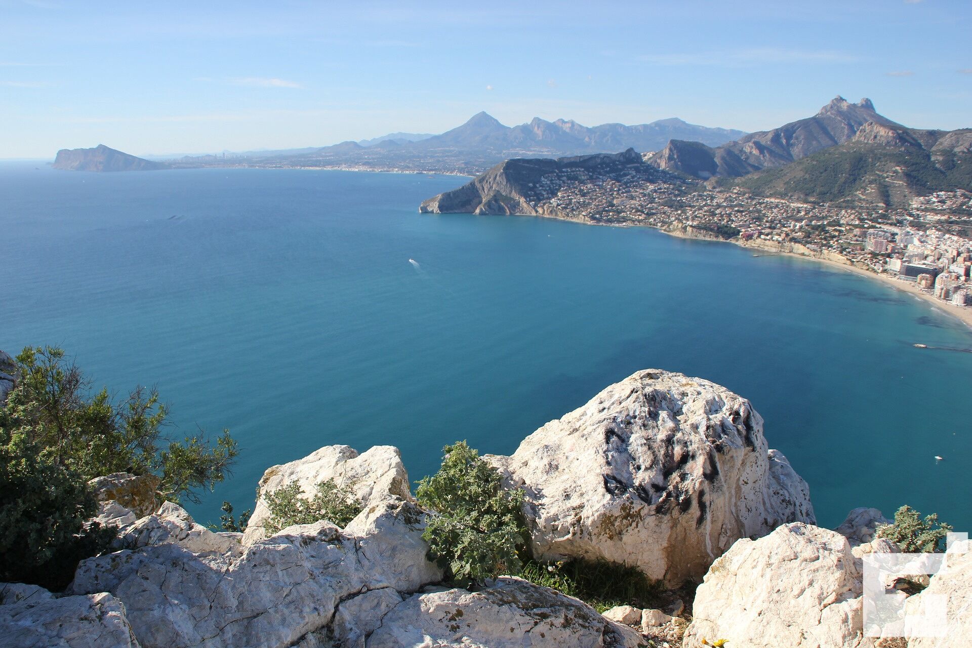 A coastal city with a prominent rocky outcrop in the foreground and a view of the city and mountains in the distance.