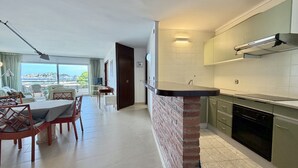 Une cuisine moderne avec un mur d’accent en briques, un coin repas avec une table et des chaises, et une vue sur la mer grâce à de grandes fenêtres.