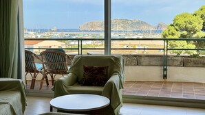 Un balcon avec vue sur un port de plaisance et une île rocheuse. On y trouve des chaises en osier et une petite table ronde.