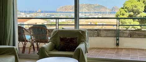 Un balcón con vista a un puerto deportivo y una isla rocosa. Hay sillas de mimbre y una pequeña mesa redonda.