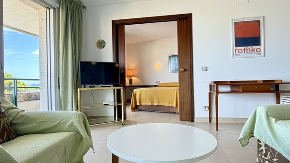 Habitación de hotel con cama, televisor sobre un mueble, una mesa y vistas al exterior.