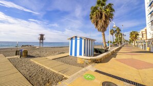 Eine Uferpromenade mit Holzsteg, einer blau-weißen Strandhütte, einem Rettungsturm und einer Palme.