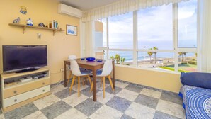 Ein Zimmer mit Fernseher, Esstisch, Stühlen und Blick auf den Strand.