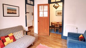 A cozy living room with a sofa, a blue armchair, and a wooden dining table with chairs. A wooden door leads to another room.