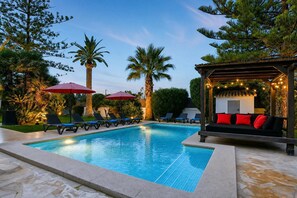 A swimming pool with lounge chairs and umbrellas, surrounded by palm trees and a wooden pergola with string lights.
