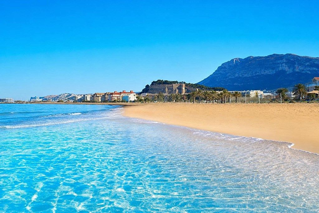 A beach with clear blue water, a sandy shore, and a mountain in the background.