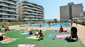 Zona de piscina con gente tomando el sol sobre esterillas, una piscina y edificios modernos al fondo.