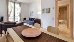 En moderne stue med sofa, spisebord med stole og et stort vindue med gardiner.