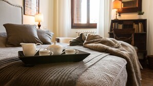 Un lit bien fait, avec un plateau contenant des tasses à thé blanches et une théière, une fenêtre avec des rideaux, une bibliothèque et une lampe.