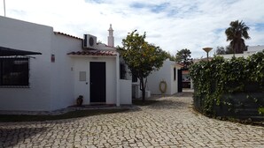 Une cour pavée de pierres, bordée de bâtiments blancs, avec une porte et un petit arbre aux fruits jaunes.