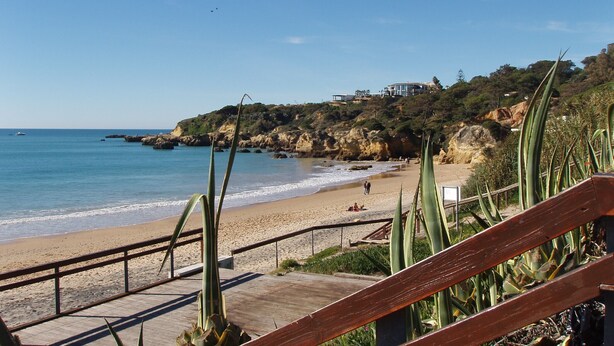 Une plage avec une promenade en bois, une falaise rocheuse et une maison au sommet.