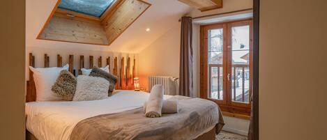 A bedroom with a sloped ceiling, a skylight, a large bed with multiple pillows, a radiator, and a view of the outside through a window.