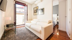Un salon moderne avec un canapé blanc, une table basse en verre et un amplificateur Marshall. Deux tableaux encadrés ornent le mur, une porte donne accès à une salle de bain, et une fenêtre offre une vue sur un bâtiment traditionnel situé à l’extérieur.