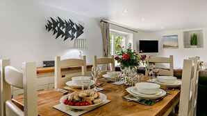 Een eetgedeelte met een houten tafel, gedekt voor vier personen, omringd door witte stoelen. Op tafel staat een vaas met bloemen, en aan de muur hangt een flatscreen televisie.