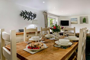 Ein Esszimmer mit einem Holztisch, gedeckt für vier Personen, weißen Stühlen, einer Vase mit Blumen und einem an der Wand befestigten Flachbildfernseher.
