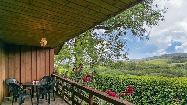 Una terraza de madera con una mesa y sillas, con vista a un exuberante jardín lleno de flores rojas y colinas ondulantes.