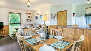 Un comedor con una mesa de madera dispuesta para cuatro personas, rodeada de sillas. Se ve un televisor al fondo y una ventana con persianas.