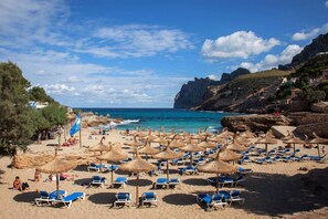 A beach with sun loungers and thatched umbrellas, rocky cliffs, and a clear blue sky.