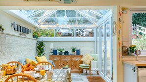 A bright kitchen with a dining area, featuring a large window, wooden cabinets, and a variety of plants. - 2 Bed in Appledore (oc-drum) (Appledore)