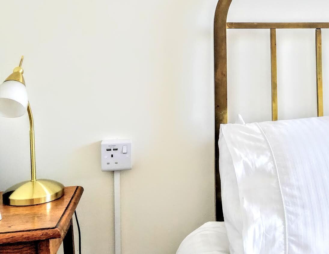 Un letto con biancheria bianca, un comodino in legno con una lampada e una presa elettrica.