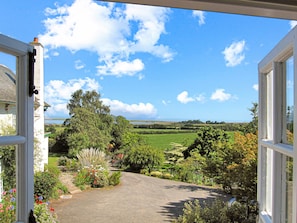 Vista a través de una puerta abierta que da a un jardín con un sendero empedrado, diversas plantas y un paisaje lejano.