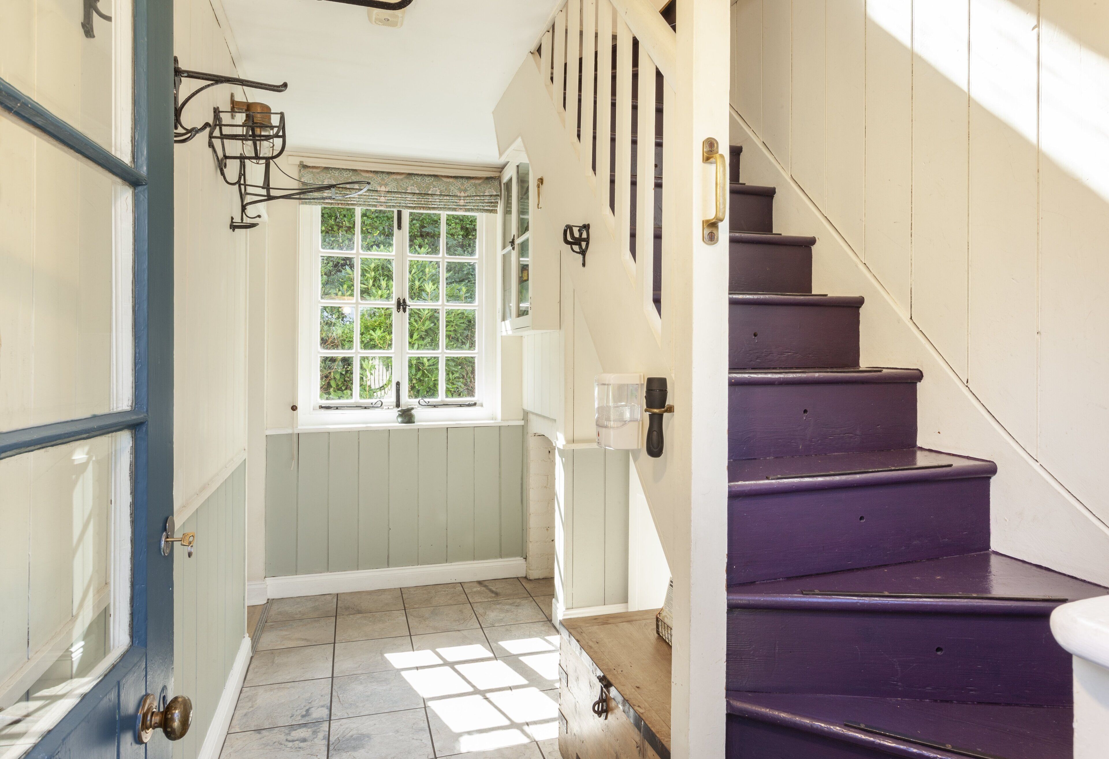 Un escalier aux marches violettes menant à un palier, une fenêtre donnant sur un paysage verdoyant et une porte dotée d’un cadre bleu.