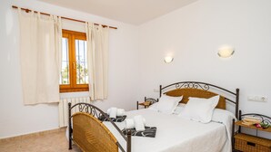 Ein ordentlich bezogenes Bett mit weißen Leinentüchern, ein Weiden-Kopfstück und ein Nachttisch mit Buch und Blumenvase.
