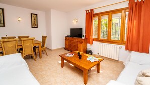 Ein möbliertes Wohnzimmer mit Holztisch, weißem Sofa, Korbstühlen, einem Fernseher und einem Fenster mit orangefarbenen Vorhängen.