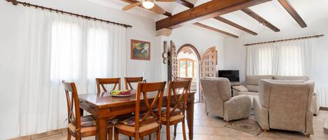 A dining area with a wooden table and chairs, a ceiling fan, and a view of the outside through an arched doorway.
