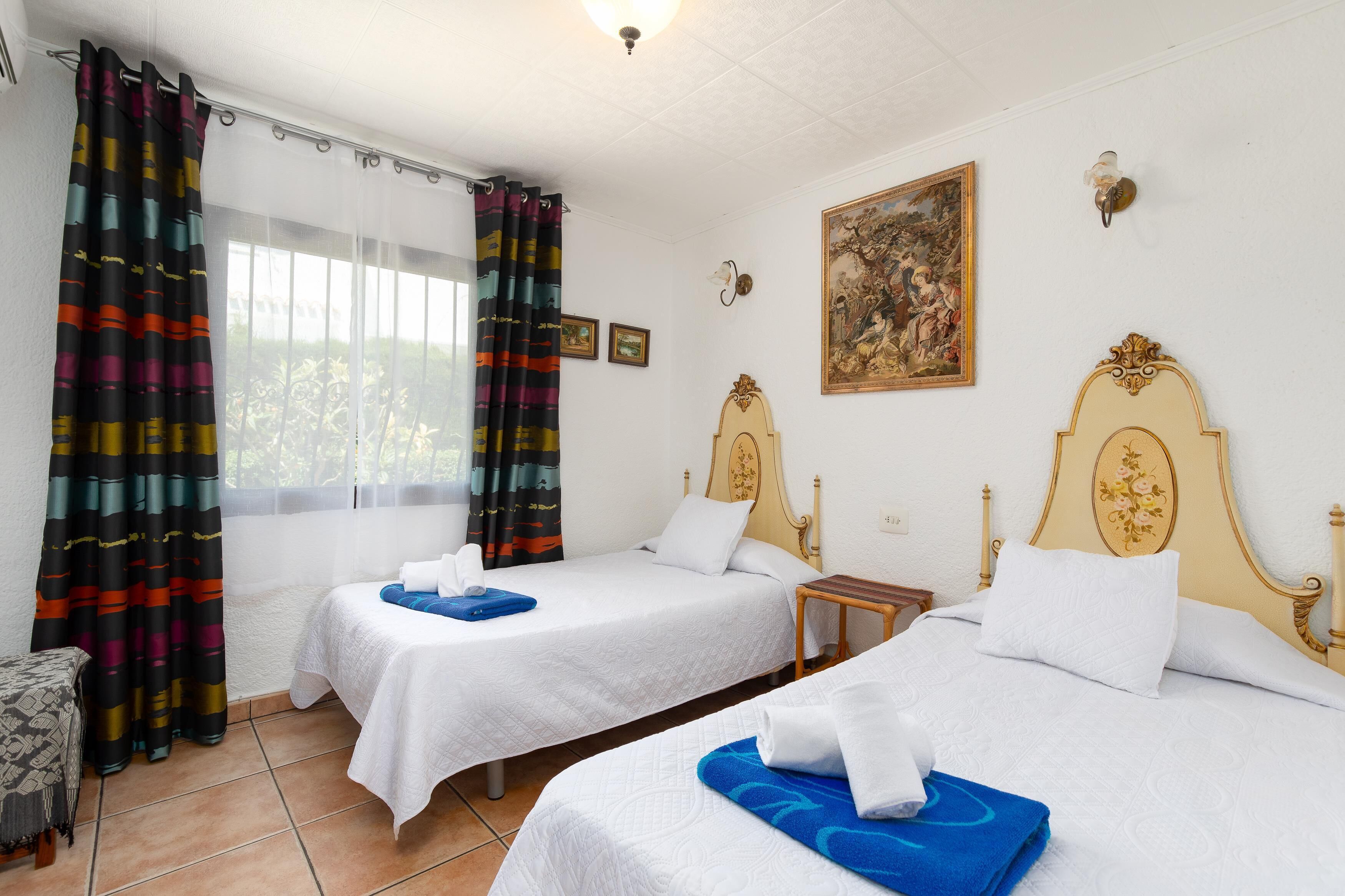 A hotel room with two beds, a window with curtains, a painting on the wall, and a small table.